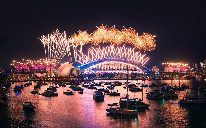 Fireworks over Sydney Harbour Bridge and Opera House at night with boats gathered on the water.