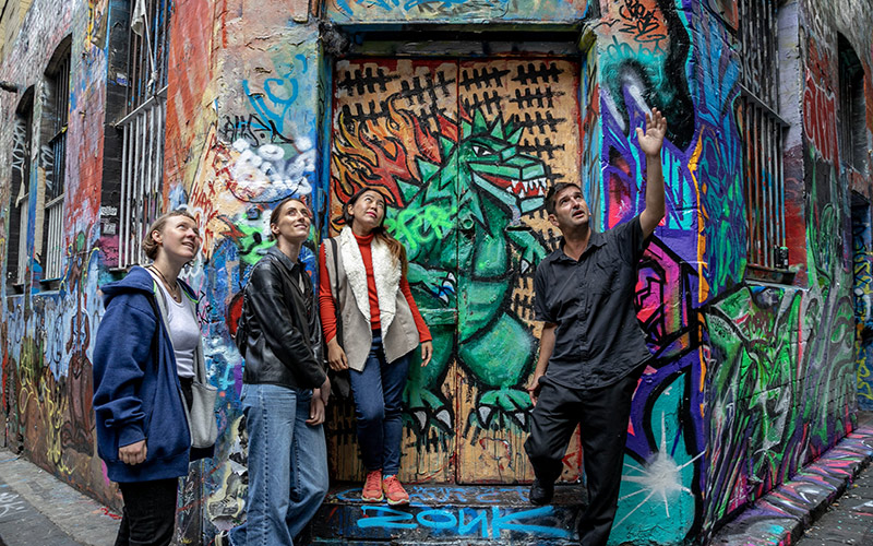 People walking through a leafy urban courtyard with tables, plants, and a large mural painted on a brick wall