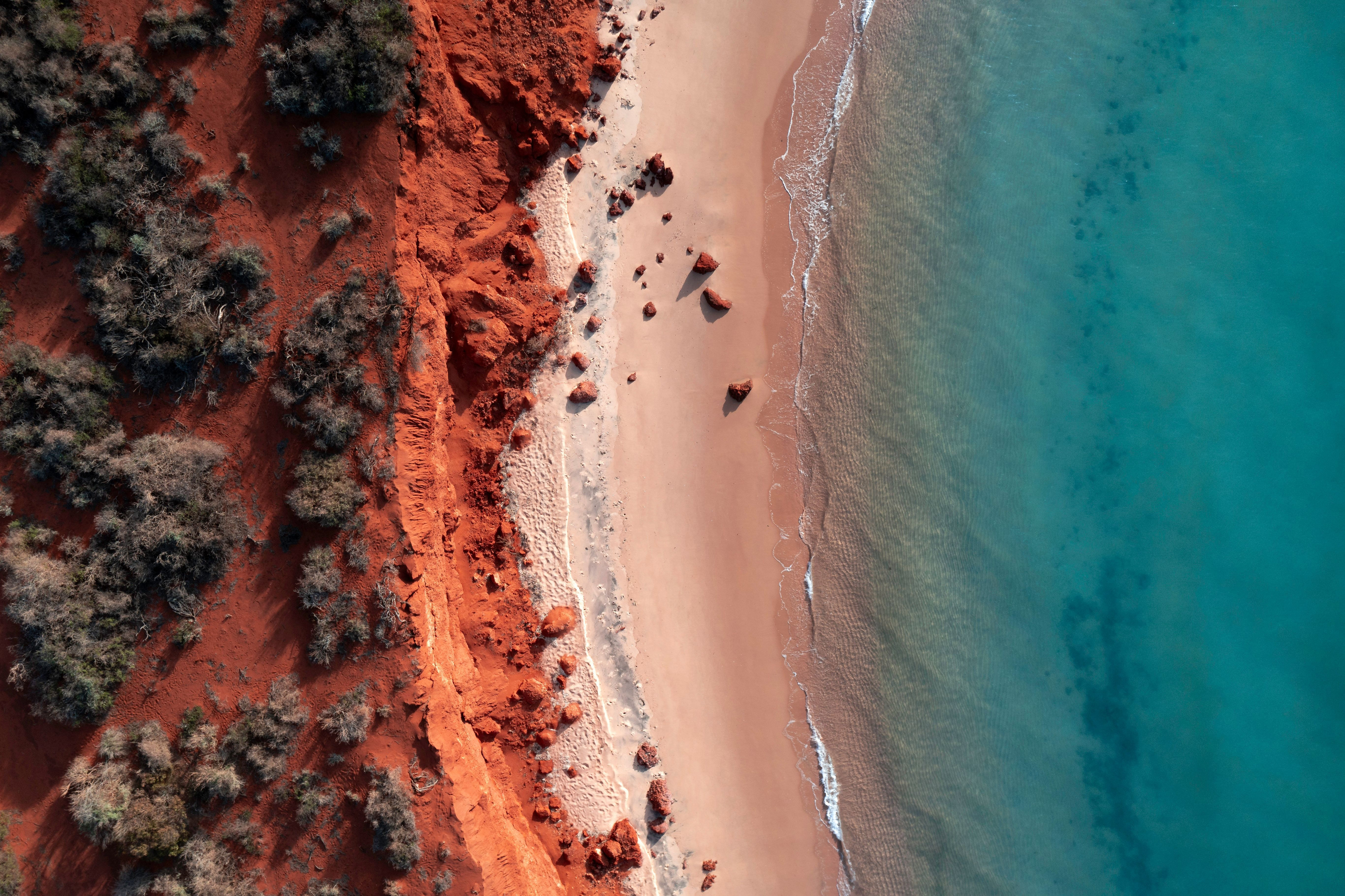 Top-down aerial view of red coastal cliffs meeting a sandy beach and calm turquoise ocean with scattered rocks.