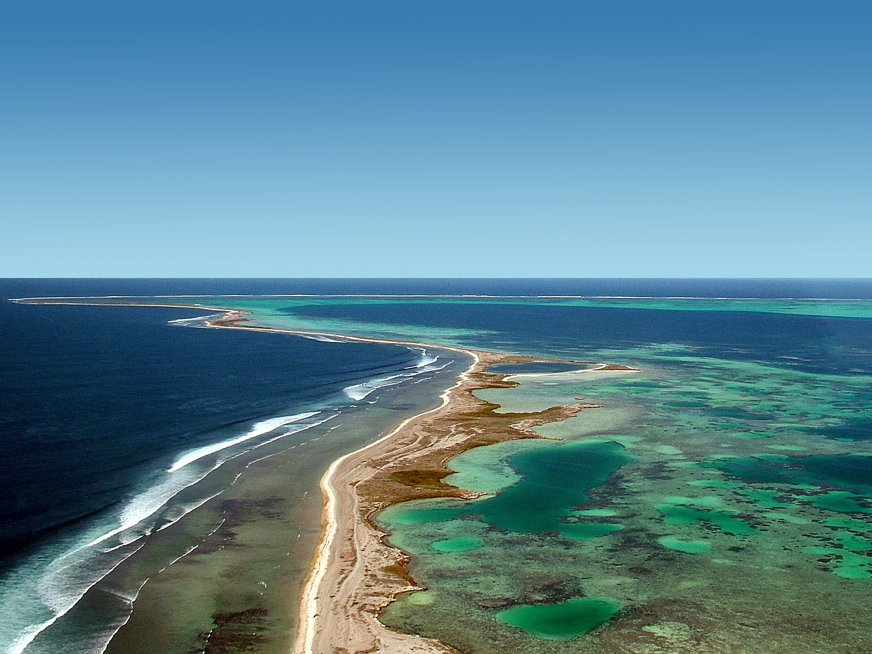 Aerial view of a long sandbar surrounded by turquoise and deep blue ocean with reef patterns and gentle waves.